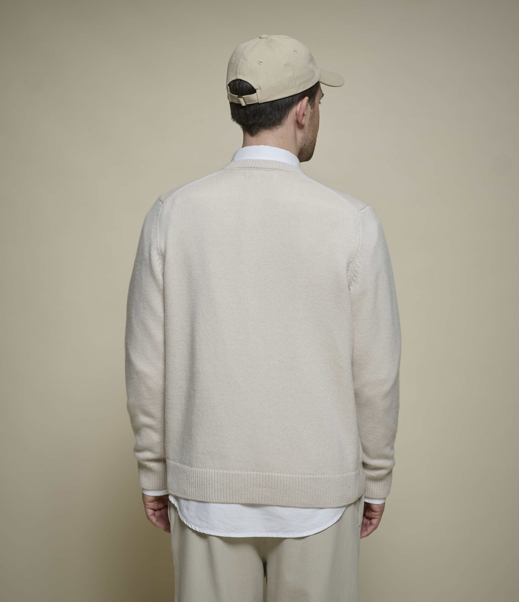 A man viewed from behind wearing a light beige knit cardigan over a white shirt, paired with beige trousers and a beige baseball cap, against a solid beige background.