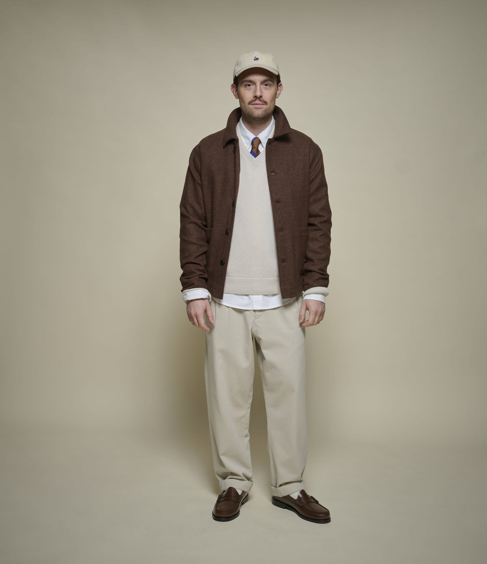 A man stands against a plain beige studio background wearing a brown overshirt over a cream sweater and white shirt, with beige trousers, brown loafers, and a cream cap.