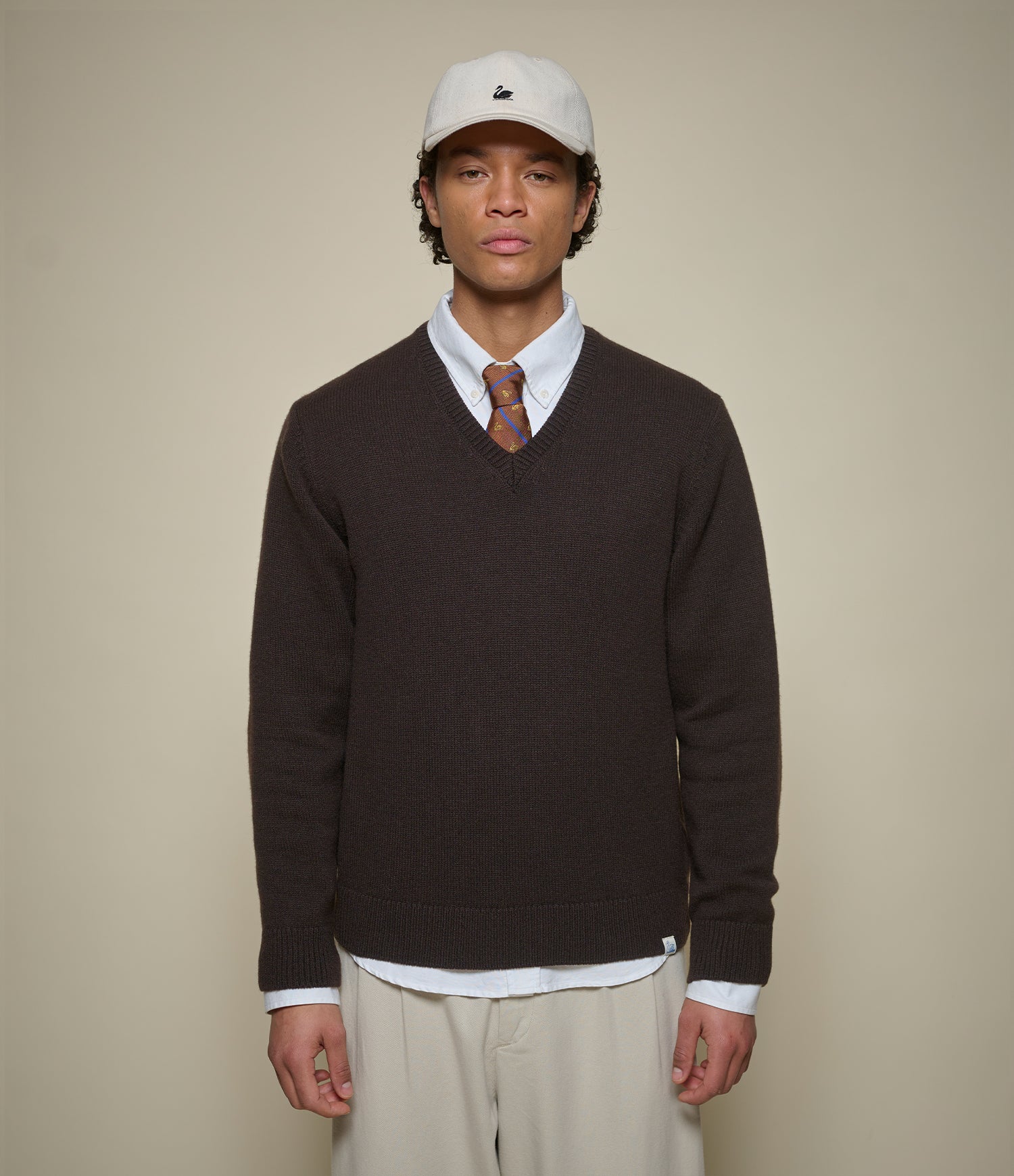 A man wearing a dark brown v-neck knit sweater over a white collared shirt and patterned tie, paired with beige trousers and a white baseball cap, standing against a solid beige background.