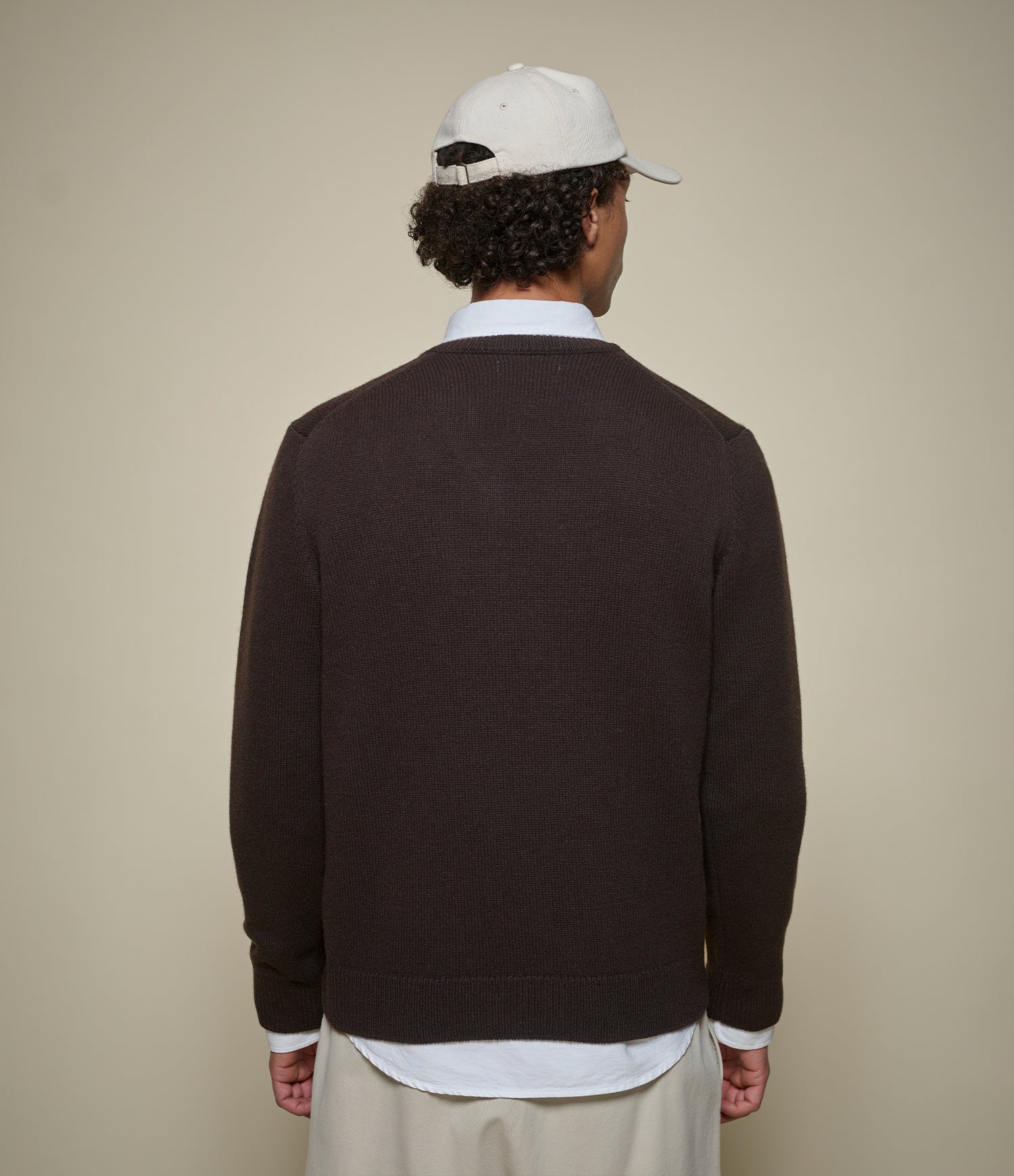 A man viewed from behind wearing a dark brown knit sweater over a white collared shirt, paired with a white baseball cap and light beige trousers, against a solid beige background.