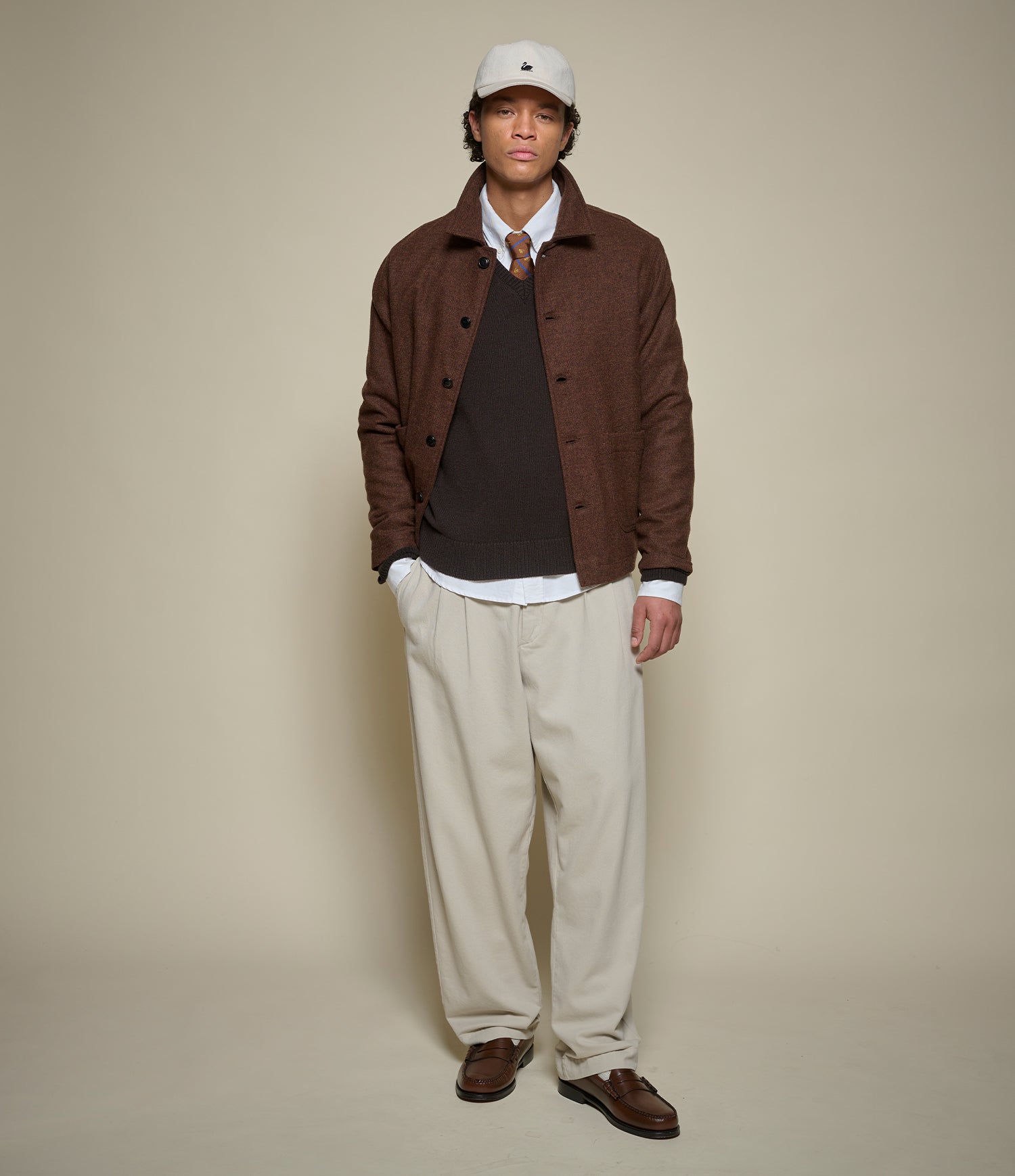 A man modeling a brown button-up overshirt over a dark brown sweater, white collared shirt, patterned tie, beige trousers, and brown loafers, against a solid beige studio background.