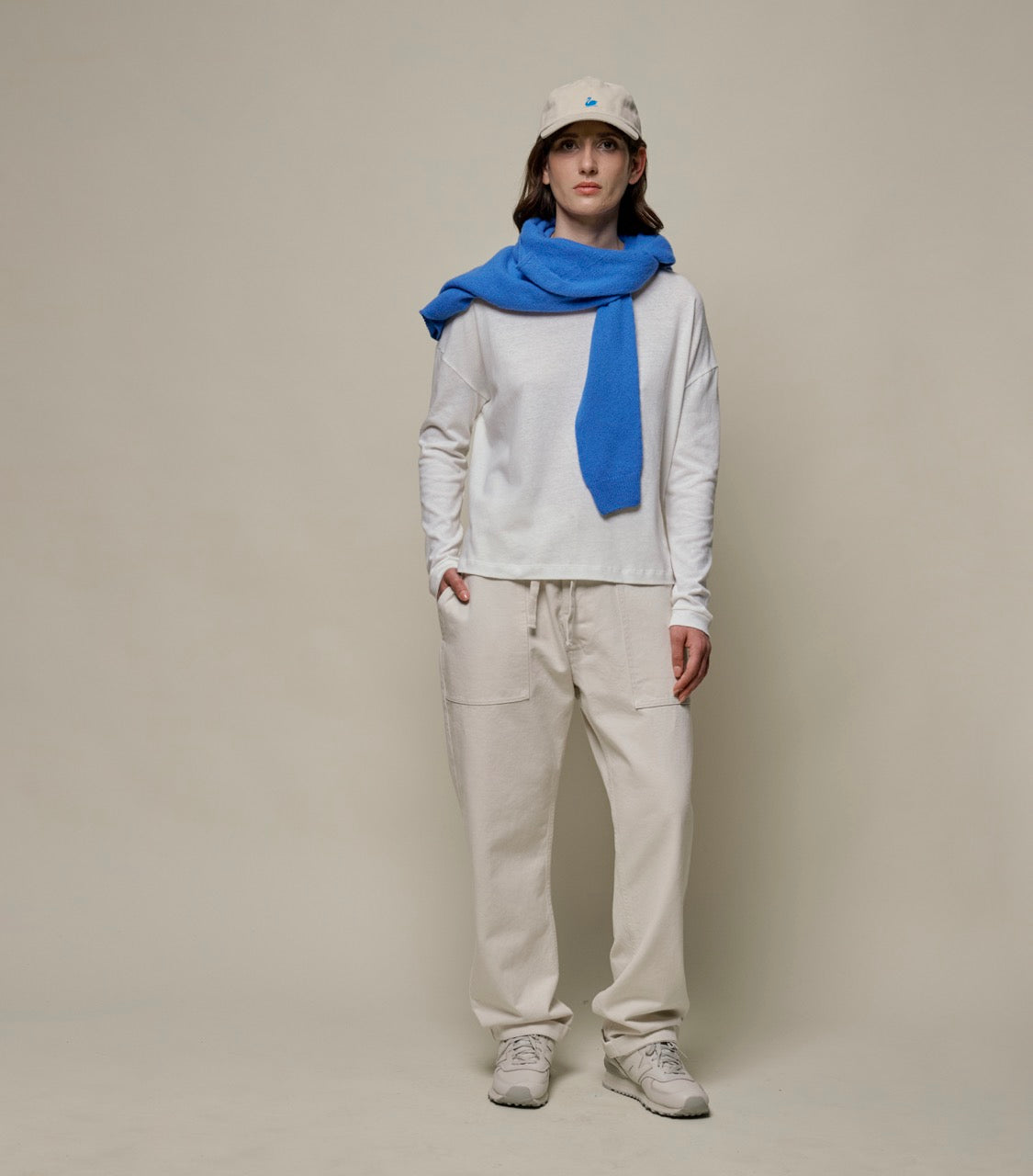 Person wearing a white long-sleeve shirt, blue scarf, beige pants, and cap on a plain background