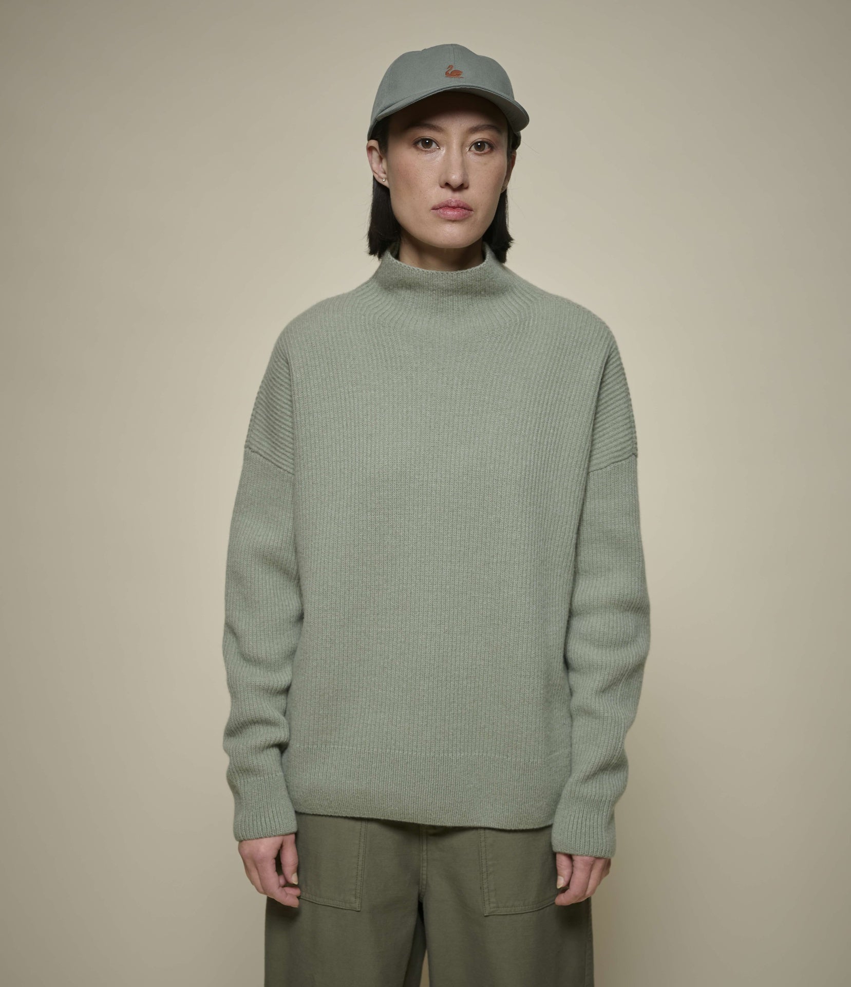 A model wearing a sage green ribbed mock neck sweater, olive green pants, and a matching sage green baseball cap, standing against a solid beige background.
