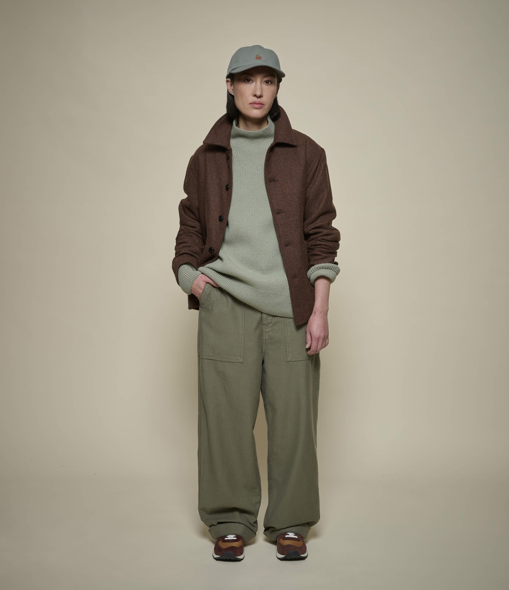 A model stands against a plain beige studio background wearing a dark brown jacket over a light sage green turtleneck sweater, olive green wide-leg pants, and a gray baseball cap.