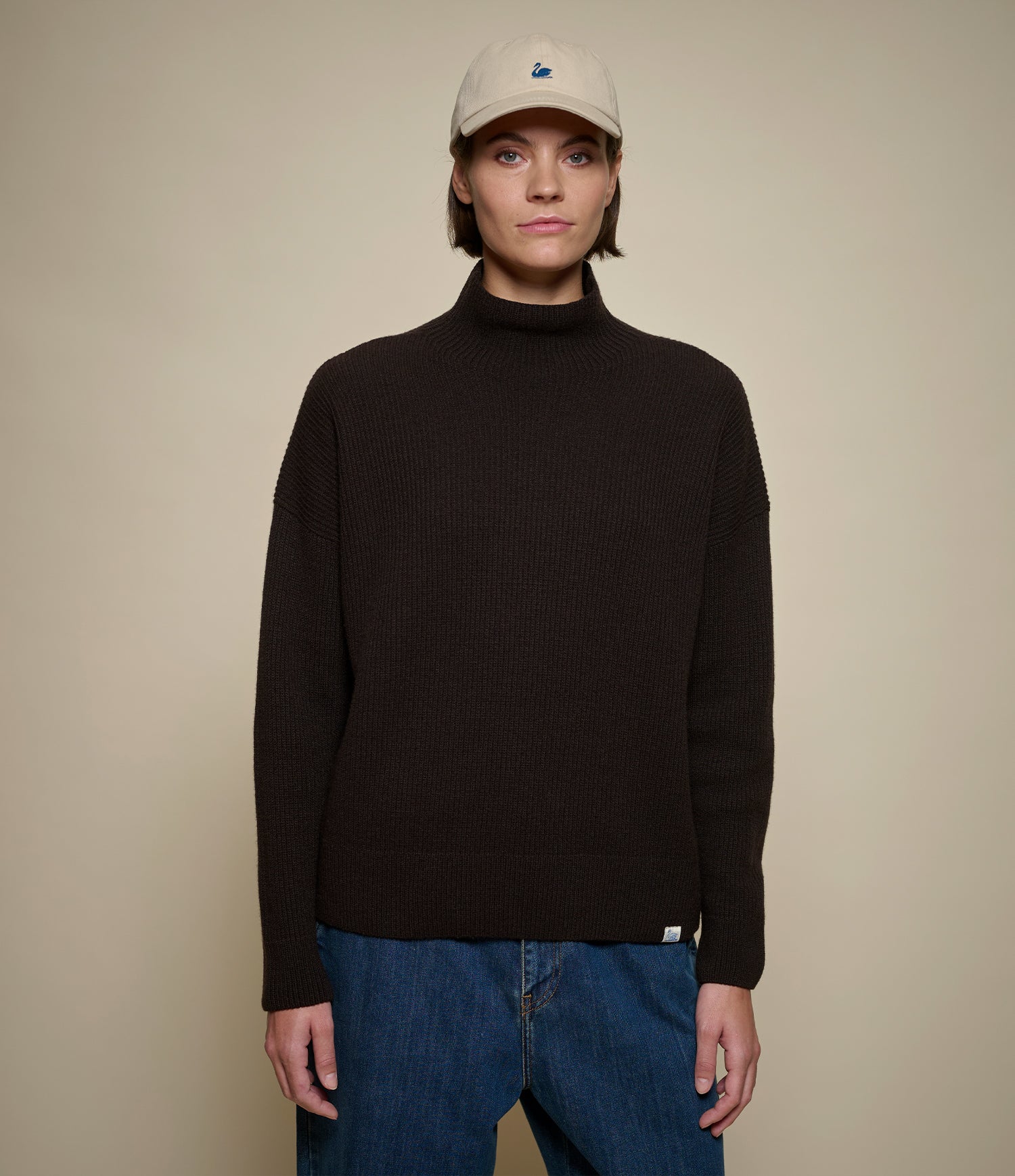 A model wearing a dark brown ribbed knit mock neck sweater, blue jeans, and a beige baseball cap, standing against a solid beige background.