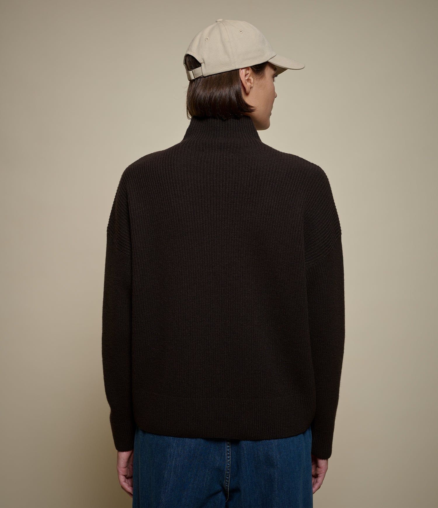 A person shown from the back wearing a dark brown ribbed turtleneck sweater, a light beige baseball cap, and blue jeans, set against a solid beige background.