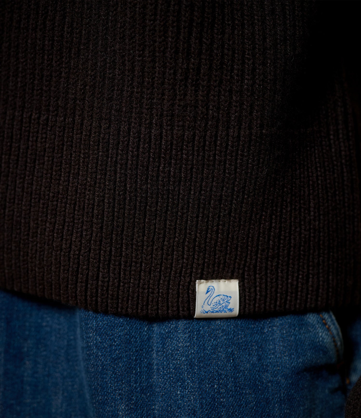 Close-up of a black ribbed knit sweater worn with blue jeans, featuring a small white woven label with a blue swan logo near the hem.