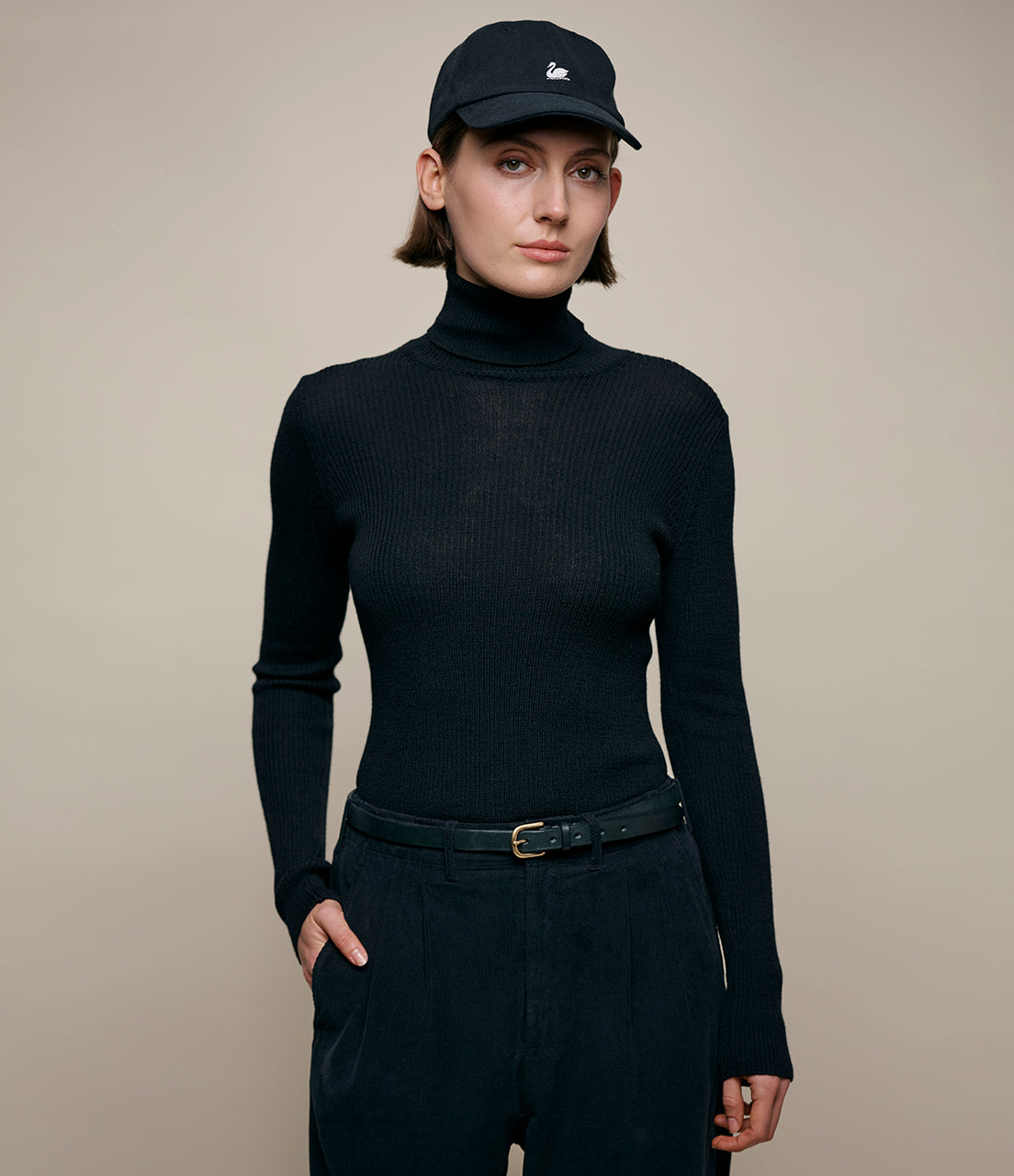 Person wearing a black turtleneck, black pants, and a black cap with a logo on a beige background