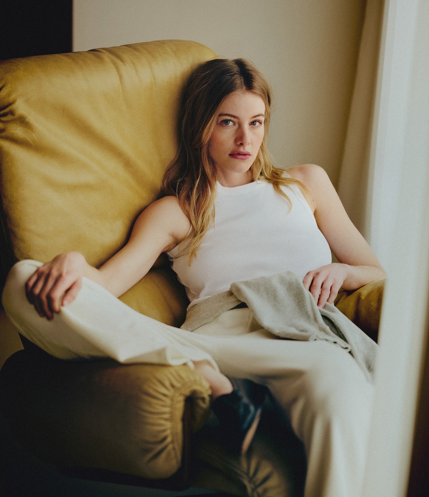 A woman reclining in a large mustard yellow armchair, wearing a white sleeveless top and light beige pants, with a light gray wrap or blanket draped across her lap.