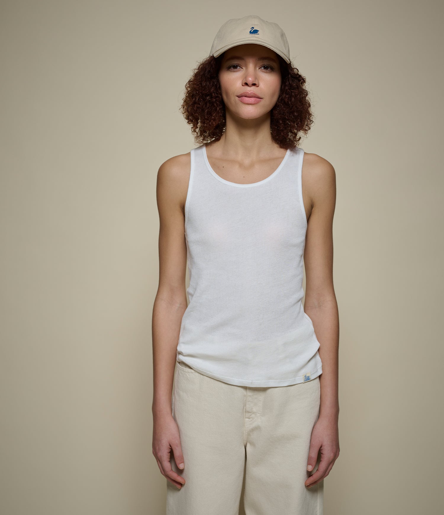 Person wearing a white ribbed tank top and beige cap against a plain background