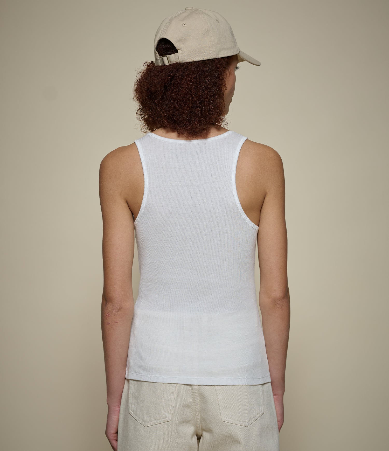 Person wearing a white ribbed tank top and beige cap on a plain background