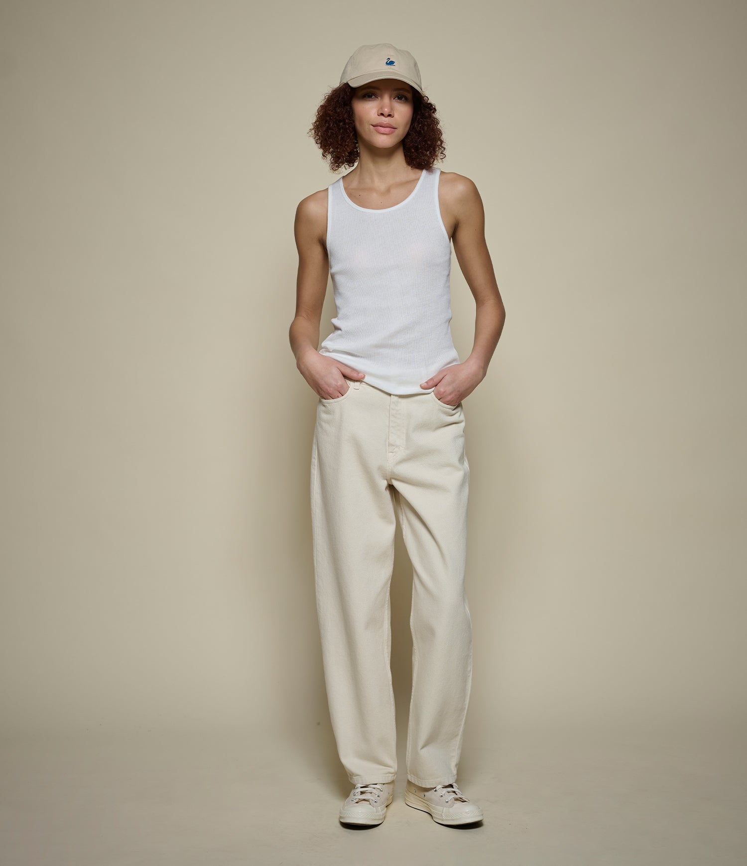 Person wearing a white tank top, beige pants, and a beige cap against a plain background