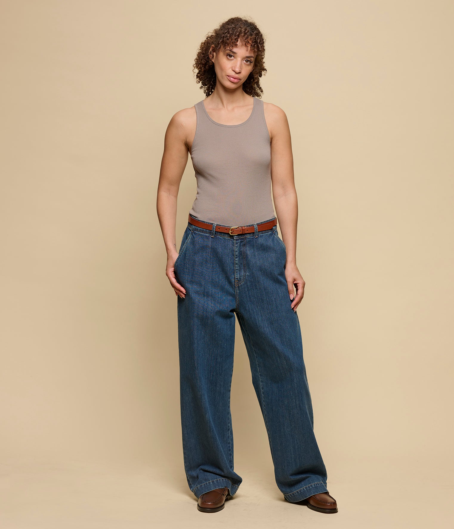 Woman wearing a brown ribbed tank top and blue jeans on a beige background