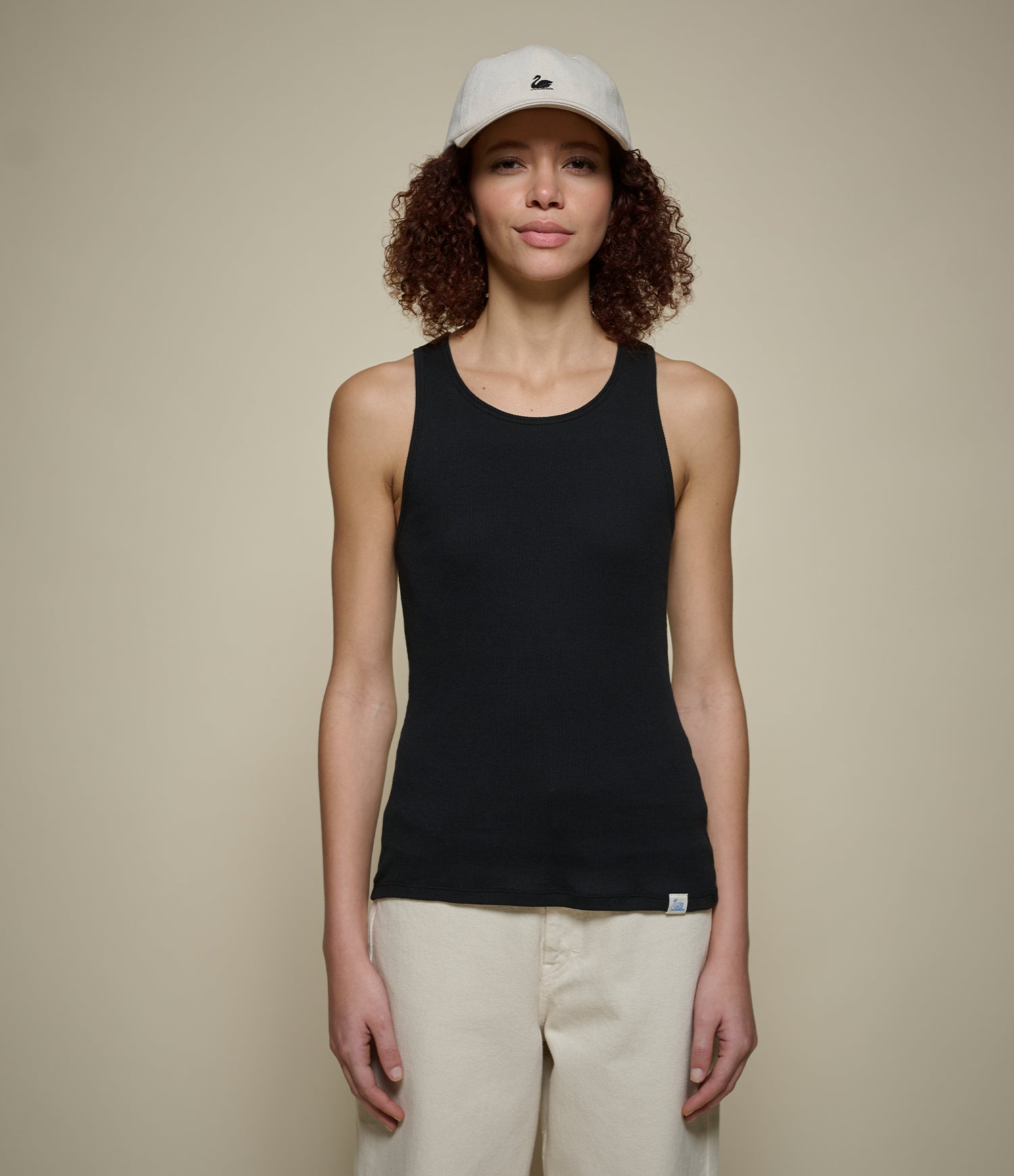 Woman wearing a black ribbed tank top and beige pants with a white cap on a beige background