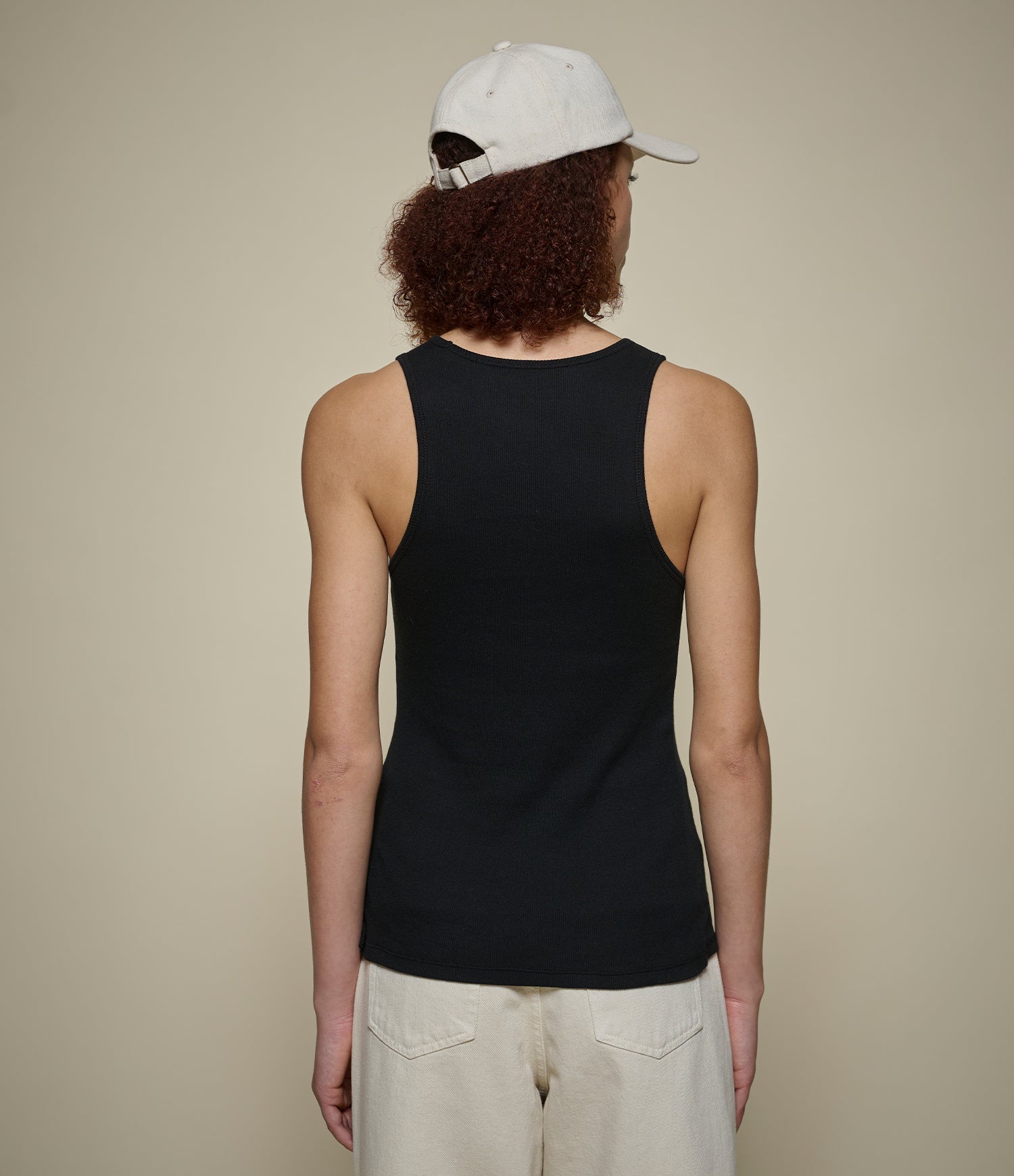 Person wearing a black ribbed tank top and beige pants with a white cap on a beige background