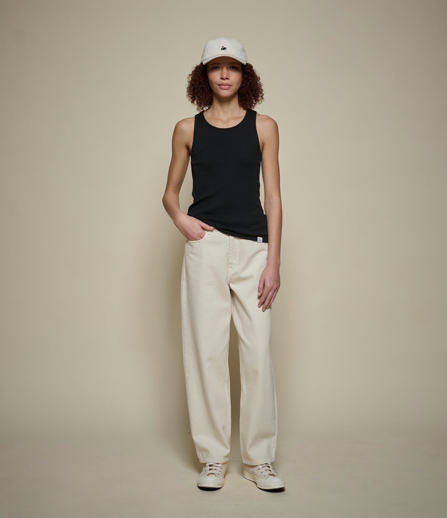 Person wearing a black tank top, beige pants, and a white cap on a beige background
