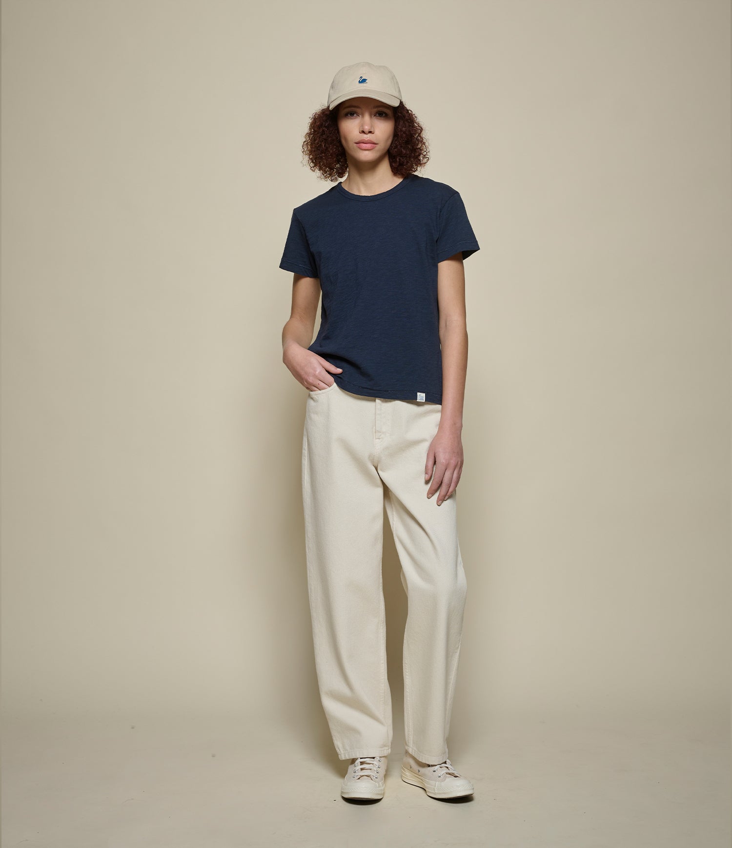 Person wearing a navy t-shirt, beige cap, and white pants against a beige background