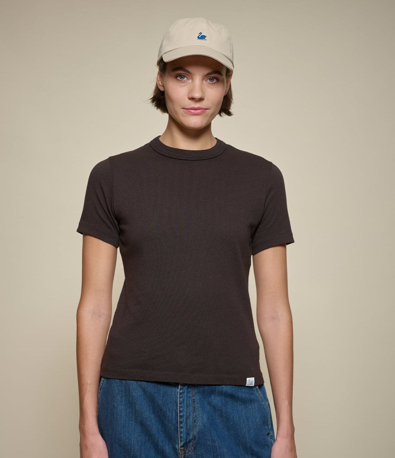 Woman wearing a dark brown short-sleeve crewneck t-shirt, blue jeans, and a beige baseball cap, standing against a solid beige background.