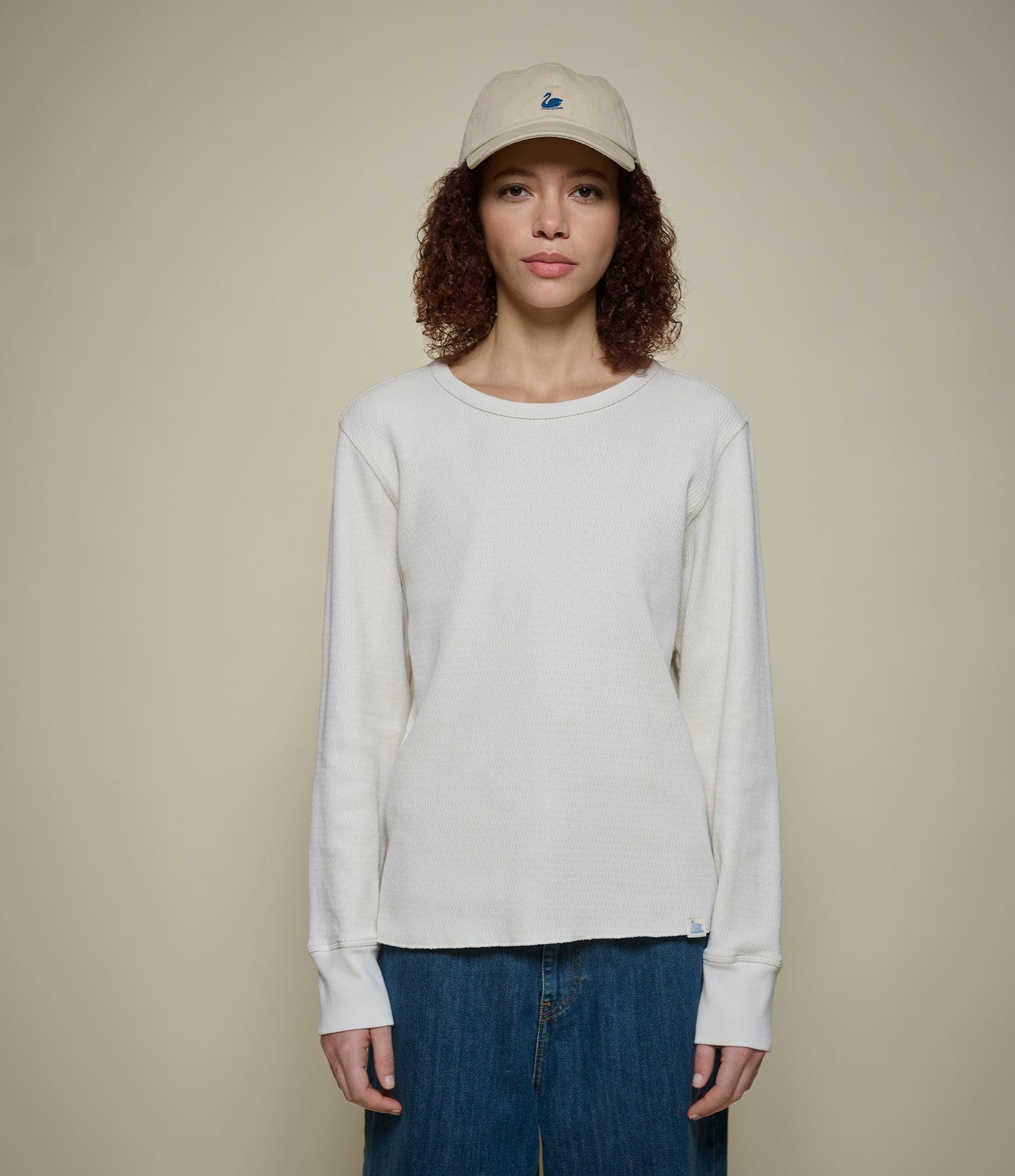 Woman wearing a white long-sleeve thermal shirt and blue jeans with a beige cap on a beige background