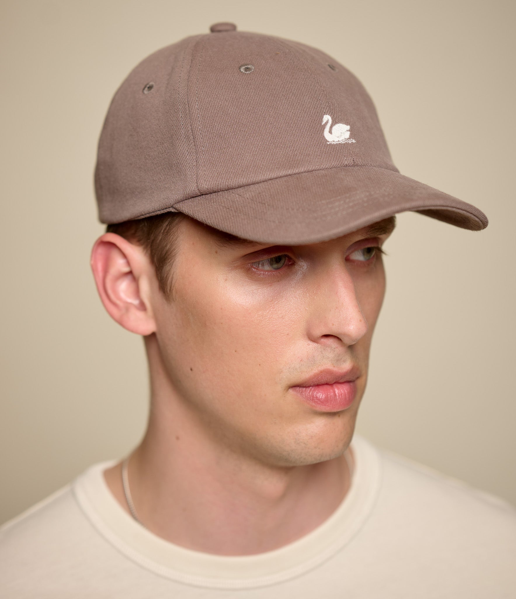 A man wearing a taupe brown baseball cap with a small white swan logo on the front, paired with a cream-colored crewneck shirt against a solid beige background.